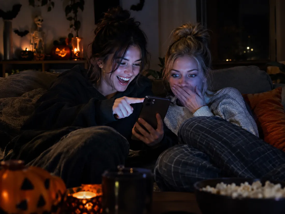 Two friends on a couch at night, one laughing at a phone while the other pretends to be scared