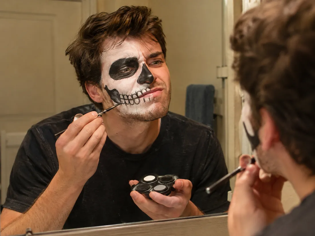 A man at a bathroom mirror struggling to paint skull face makeup with a brush