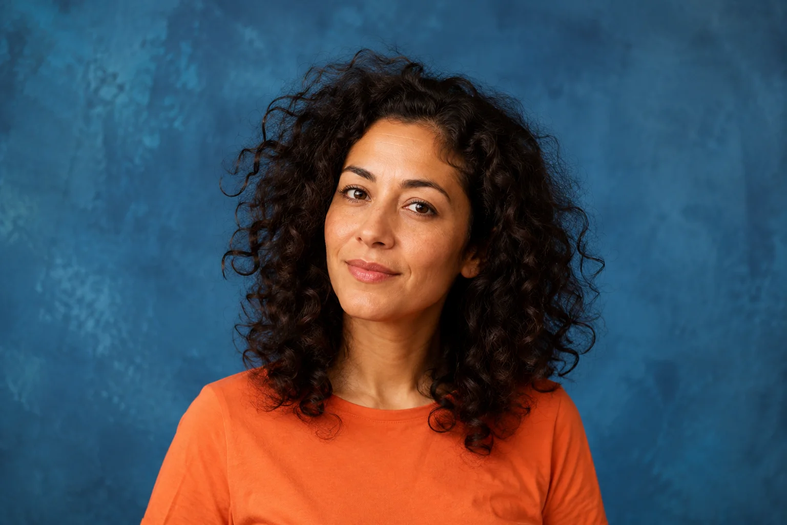 Woman with curly dark hair and an orange shirt against a blue studio backdrop